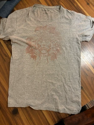 Grå t-shirt med lejontryck  Denim & Supply Ralph Lauren - Snygg grå t-shirt från Denim & Supply Ralph Lauren med ett diskret lejontryck framtill. Tillverkad i mjuk bomullsblandning (90% bomull, 10% polyester) och har klassisk passform med korta ärmar och rund hals.