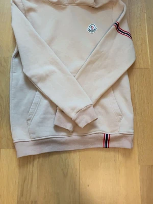 Beige hoodie från Moncler - Snygg beige hoodie från Moncler med klassisk huva och fickor framtill. Har Moncler-logga på bröstet och coola röd-blå-vita ränder på ena ärmen och vid nederkanten. Tillverkad i mjukt bomullsmaterial som känns skönt mot huden. Perfekt för en avslappnad och trendig look.