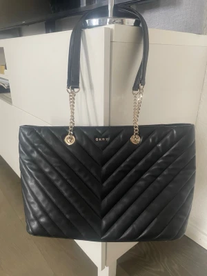 Svart quiltad handväska från DKNY - Snygg svart handväska från DKNY i quiltat mönster med guldfärgade kedjedetaljer och handtag i läder. Väskan har en stilren, rektangulär form och diskret DKNY-logga framtill. Perfekt för dig som gillar klassisk men trendig design.