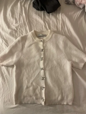 Vit kortärmad tröja från Zara - Supersöt vit tröja från Zara med korta ärmar och ribbade kanter. Koftan har fina dekorativa knappar framtill som ger en snygg och unik touch. Mjuk och skön kvalitet. 