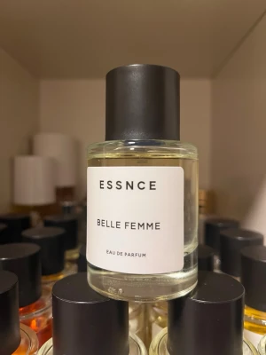 ESSNCE Belle Femme Eau de Parfum - Minimalistisk parfymflaska från ESSNCE med doften Belle Femme. Flaskan är cylindrisk i genomskinligt glas med svart rund kork och vit etikett med svart text. En stilren och modern parfym som passar perfekt i badrumshyllan.
