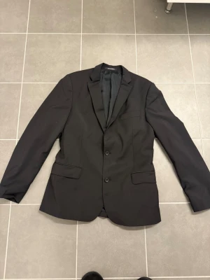 Svart kavaj från Oscar Jacobsson - Stilren svart kavaj från Oscar Jacobsson med två knappar framtill, klassisk krage och tre fickor. Kavajen har en rak passform och är helfodrad för extra komfort. Perfekt för dig som vill ha en enkel och snygg look till vardags eller mer uppklädda tillfällen.