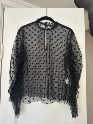 Svart meshblus med prickar Topshop - Säljer en transparent svart meshblus från Topshop med stora sammetsprickar. Blusen har långa ärmar, voluminös passform och knytning i nacken. Perfekt för att styla lager på lager och ger en trendig look.