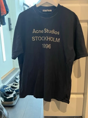 Svart Acne Studios t-shirt Stockholm - Svart t-shirt från Acne Studios med tryck 'Acne Studios STOCKHOLM 1996' på bröstet. Klassisk passform och rund hals. Perfekt för dig som gillar stilrena och trendiga plagg med streetkänsla.