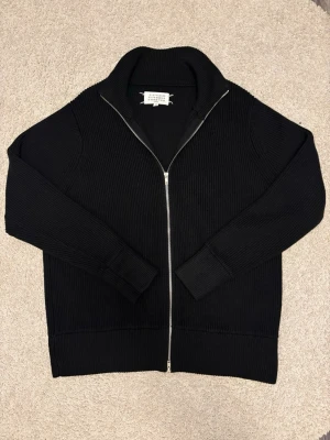 Svart stickad ziptröja M M( cop) - Svart stickad ziptröja med hög krage och hel dragkedja framtill. Tröjan har ribbstickad struktur och diskreta vita sömmar bak i nacken, klassiskt för märket. Perfekt för lager-på-lager och stilren look. Passar som L-XL