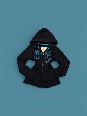 Svart hoodie från Hollister - Svart hoodie med dragkedja från Hollister. Stor blå text 'Hollister California' på bröstet och klassisk logga. Mjuk bomullsblandning, två fickor fram och huva. Perfekt för en avslappnad och cool stil. Storlek S