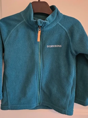 Mörkgön, turkoz barnfleecejacka från Didriksons str 90 - Mysig blå fleecejacka från Didriksons med dragkedja framtill och hög krage. Jackan har en enkel och sportig design, perfekt för lager-på-lager och chill dagar. Materialet är mjukt och värmande fleece.