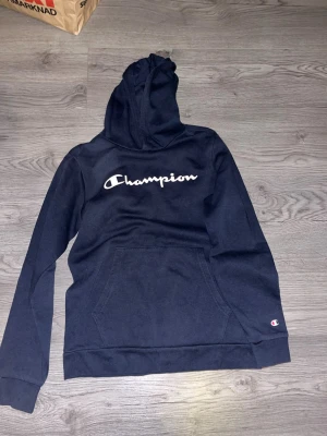 Mörkblå Champion hoodie med huva - Snygg mörkblå hoodie från Champion med stor logga i vitt över bröstet och klassisk känguruficka framtill. Hoodien har en huva och ribbade muddar vid ärmar och nederkant. Perfekt för en avslappnad och sportig stil. Storlek-13/14 years old/162-167 cm