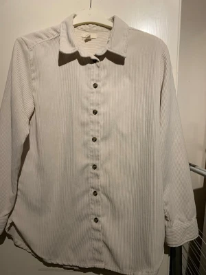 Beige overshirt i manchester - Stilren beige overshirt i mjuk manchester med klassisk krage och knappar framtill. Overshirten har långa ärmar och en avslappnad passform, perfekt att slänga över en t-shirt. Materialet är ribbat och ger en trendig look.