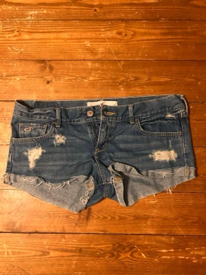 Blå jeansshorts från Hollister - Säljer ett par blå jeansshorts från Hollister med slitna detaljer och råa kanter. Klassisk femficksmodell med låg midja och knappgylf. Perfekta för sommaren och har en avslappnad, trendig look. (Pris kan diskuteras💕)