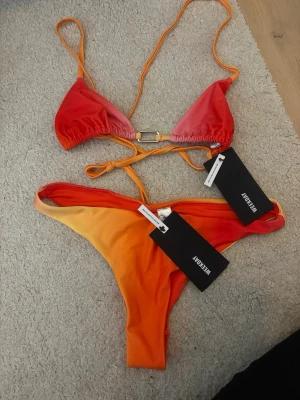 Orange och röd bikini från Weekday - Snygg bikini från Weekday i starka orangea och röda nyanser. Överdelen har trekantsform med smala band och metallspänne, underdelen är enkel med hög skärning. Tillverkad i återvunnen polyester. Perfekt för stranden eller poolen!