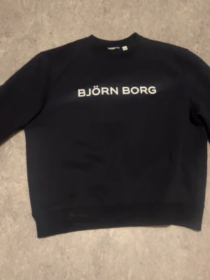Mörkl blå sweatshirt från Björn Borg - En mörkblå sweatshirt från Björn Borg med vit logga tryckt över bröstet. Tröjan har rund halsringning och långa ärmar. Tillverkad i mjukt material som känns skönt mot huden. Perfekt för en avslappnad och sportig look.