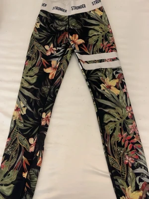 Stronger leggings med blommigt mönster - Snygga leggings från Stronger med tropiskt blommönster i grönt, orange och rosa. Bred vit resår i midjan med svart logga och tre vita ränder på ena benet. Perfekta för träning eller chill. Materialet är stretchigt och följsamt.