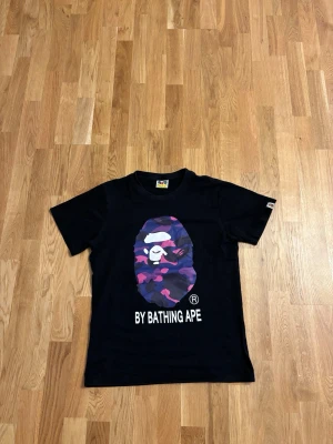 Svart BAPE t-shirt med lila camo-print - Säljer en svart t-shirt från By Bathing Ape med det ikoniska BAPE-apans ansikte i lila, rosa och blått camo-print på bröstet. T-shirten har korta ärmar, rund hals och trycket 'BY BATHING APE' under motivet. Perfekt för dig som gillar streetwear.
