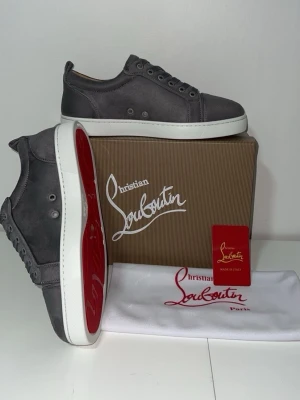 Gråa sneakers från Christian Louboutin - Snygga gråa sneakers från Christian Louboutin med klassisk röd sula och vita kanter. Skorna har snörning och är tillverkade i mocka för en lyxig känsla. Perfekta för dig som vill sticka ut med stilrena detaljer och exklusivt märke.