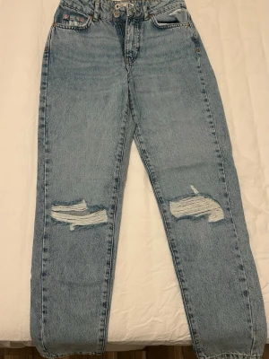 Ljusa mom jeans med slitningar från Gina Tricot  - Ett par ljusblå mom jeans med raka ben från Gina Tricot i storlek 32. Jeansen har tydliga slitningar över knäna och klassiska fem fickor. Aldrig använda.