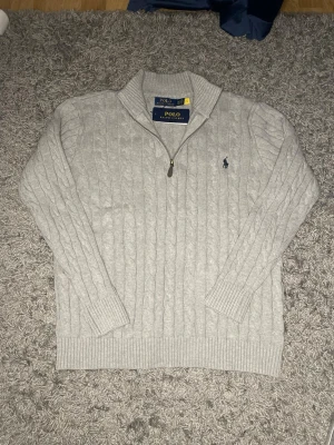 Ljusgrå kabelstickad tröja Polo Ralph Lauren - Snygg ljusgrå kabelstickad tröja från Polo Ralph Lauren med halv dragkedja och ribbad krage. Tröjan har långa ärmar, ribbade muddar och den klassiska broderade loggan på bröstet. Perfekt för lager på lager och stilren look.