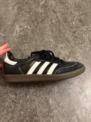 Adidas Samba svarta sneakers - Klassiska Adidas Samba sneakers i svart läder med vita detaljer och tre vita ränder på sidorna. Skon har svart mocka vid tån, svart snörning och en brun gummisula med mönster undertill. Perfekt för dig som gillar retrostil och streetwear. Som nya 