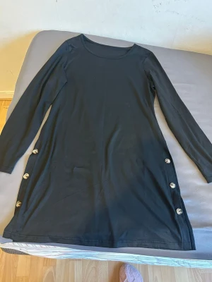 Svart långärmad klänning med knappar - En svart långärmad klänning med rund halsringning och dekorativa knappar längs sidorna. Klänningen har en enkel och stilren design som passar perfekt till många olika tillfällen.