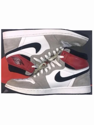 Nike Air Jordan 1 Retro High OG - Nike Air Jordan 1 Retro High OG med vit läderbas, grå mockadetaljer och röda samt svarta partier runt ankeln. Klassisk svart swoosh på sidan och vita skosnören. Ikonisk high-top siluett som sticker ut. OG Box. Givetvis äkta. Några flaws se bilder. 