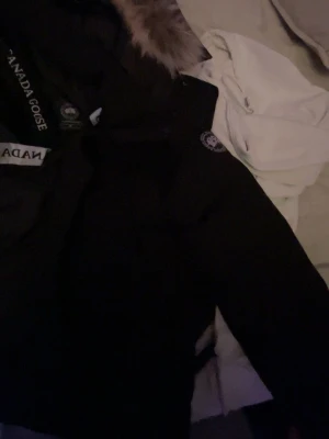 Svart parkas från Canada Goose - Säljer en svart parkas från Canada Goose med klassisk pälsdetalj på huvan och tydlig logotyp på ärmen. Jackan har en rak passform och är designad för att hålla värmen under kalla dagar. Perfekt för dig som vill ha både stil och funktion. Pälsen går även sätta på och ta av.