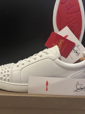 Vita sneakers från Christian Louboutin - Säljer ett par vita sneakers från Christian Louboutin med klassisk röd sula och coola nitar framtill. Skorna är låga och har en stilren design i slätt läder med vita snören. Perfekta för dig som vill sticka ut med exklusiva detaljer.