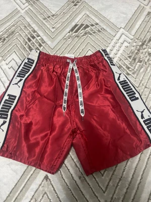Röda shorts från Puma med snörning - Röda shorts från Puma med vita sidor där det står PUMA i svart. Shortsen har elastisk midja och vit snörning med svart logga. Materialet är glansigt och känns sportigt, perfekt för en aktiv stil. Bad shorts, lite sönder på mitten men inga fel