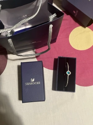 Swarovski armband med blå kristall - Elegant armband från Swarovski med en gnistrande blå kristall i mitten, omgiven av små klara stenar. Armbandet är silverfärgat och har en tunn, stilren design med justerbar kedja. Perfekt för dig som gillar exklusiva accessoarer med lite extra bling.