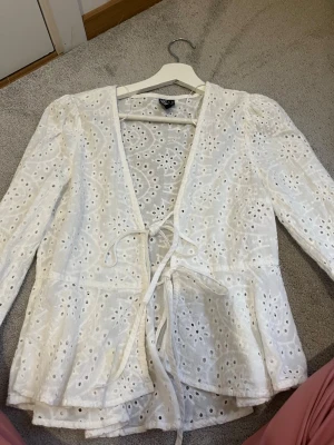 Vit broderad blus med knytband - Säljer en söt vit blus från Pieces med broderade hålmönster och volangkant. Blusen har långa ärmar, v-ringning och knytband framtill. Perfekt för en fräsch och somrig look!