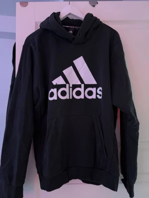 Svart hoodie från Adidas med logga - Svart hoodie från Adidas med stor vit logga och text på bröstet. Klassisk känguruficka framtill och justerbar huva. Tillverkad i mjukt bomullsmaterial som är skönt att ha på sig. Perfekt för en sportig och avslappnad stil.