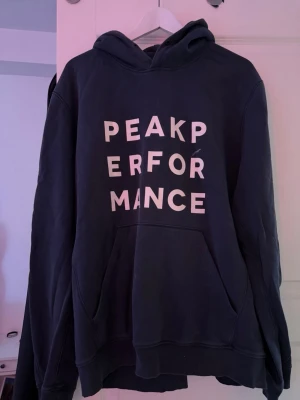 Svart hoodie från Peak Performance - Snygg svart hoodie från Peak Performance med stor vit text på bröstet. Klassisk känguruficka framtill och huva med dragsko. Perfekt för en avslappnad och sportig stil. Tillverkad i mjukt bomullsmaterial som känns skönt mot huden.