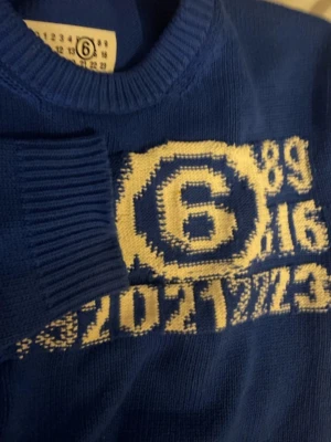 Blå stickad tröja Maison Margiela - Cool blå stickad tröja från Maison Margiela med gul grafik och siffror på bröstet. Tröjan har ribbade muddar och rund hals. Perfekt för dig som gillar streetwear och unika detaljer.