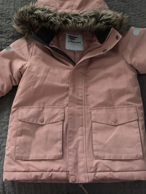 Barnjacka Rosa parkasjacka från Nordbjørn - Storlek 134 Ljusrosa parkasjacka från Nordbjørn med stor huva och fuskpäls. Jackan har två stora fickor med lock framtill, reflexdetaljer på ärmen och tryckknappar. Materialet är tåligt och vattenavvisande, perfekt för kalla dagar.