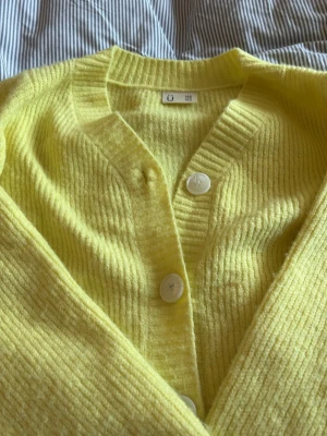 Gul ribbstickad kofta med knappar - Snygg gul ribbstickad kofta med stora vita knappar framtill och bred ribbad kant vid halsen. Går perfekt med ett linne under till eller för sig sjäkv. Koftan har en avslappnad passform och är perfekt för lager på lager. Materialet är mjukt och mysigt, perfekt för kyligare dagar men går lika bra på en kyligare sommarkväll med t.ex. kjol till! Storleken fungerar även till XS
