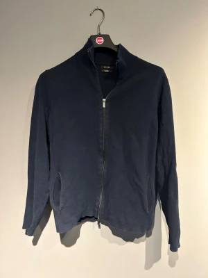 Mörkblå ziptröja från Massimo Dutti - Mörkblå ziptröja från Massimo Dutti med hög krage och två fickor framtill. Tröjan har en stilren design med långa ärmar och dragkedja hela vägen. Perfekt för dig som gillar en enkel men snygg look. Storlek L men sitter som M