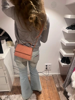 Rosa axelväska från Åhléns  - Säljer en snygg rosa axelväska med croco-mönstrad yta och justerbar axelrem. Väskan har en stilren, rektangulär form och stängs med lock. Perfekt för dig som vill ha en trendig accessoar som sticker ut.