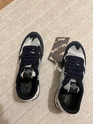 Valentino Garavani svarta och vita sneakers - Snygga sneakers från Valentino Garavani i svart och vitt med mesh- och läderdetaljer. Skorna har svarta snören, logga på plösen och en cool, sportig design. Perfekta för dig som gillar exklusiva märken och streetstyle.