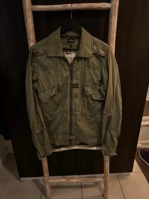 Grön vindjacka från G-Star RAW - Snygg militärgrön vindjacka från G-Star RAW med dragkedja framtill, stora fickor med knappar och coola detaljer på ärmarna. Jackan har klassisk krage och är perfekt för dig som gillar streetwear och utilitystil.