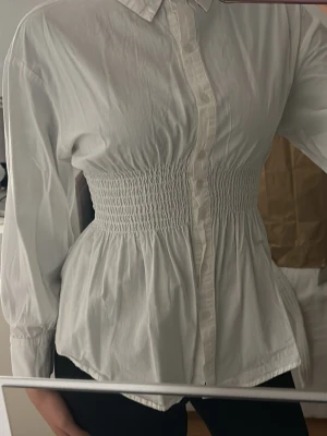 Vit blus med smockad midja - Stilren vit blus med krage och knäppning framtill. Blusen har långärmad design och smockad midja som ger en snygg siluett. Tillverkad i ett lätt bomullsliknande material som känns fräscht och passar till många olika looks.