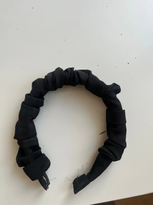 Svart rynkat diadem i tyg - Säljer ett svart diadem med rynkad design. Diademet är klätt i tyg och har en bred, puffig form som ger en trendig look. Perfekt för att lyfta vilken outfit som helst med en enkel accessoar.