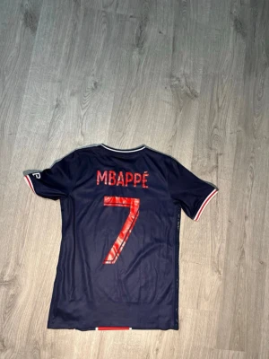 PSG matchtröja Mbappé #7 Nike - Snygg mörkblå PSG matchtröja från Nike med Mbappé och nummer 7 på ryggen. Tröjan har röda och vita detaljer, klubbmärke och sponsortryck på bröstet samt korta ärmar. Perfekt för dig som älskar fotboll och vill stötta Paris Saint-Germain.