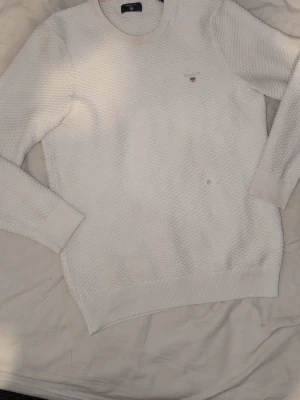 Vit GANT tröja - Vit långärmad tröja från GANT med diskret strukturmönster och rund halsringning. Liten broderad logotyp på bröstet. Tröjan har ribbade muddar vid ärmslut och nederkant. Perfekt för en clean och klassisk stil. Har små fläckar här och där och ett litet hål på magen 