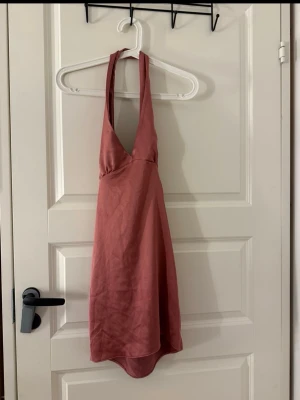 Rosa omlottklänning med knyt i satin - Snygg kort rosa klänning i satin med djup v-ringning och knyt i ryggen. Klänningen är ärmlös och har en lätt glansig finish.