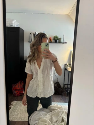 Vit blus med spetsdetaljer - Söt vit blus med korta ärmar och v-ringning. Blusen har dekorativa spetsdetaljer längs knäppningen och ett lätt transparent tyg med struktur. Perfekt att styla med jeans eller kjol för en trendig look.