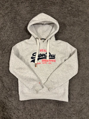 Ljusgrå hoodie från Superdry - Ljusgrå hoodie från Superdry med tryck i marinblått och rosa på bröstet. Klassisk huva med vita dragsnören. Osäker på storleken men den sitter som XS.