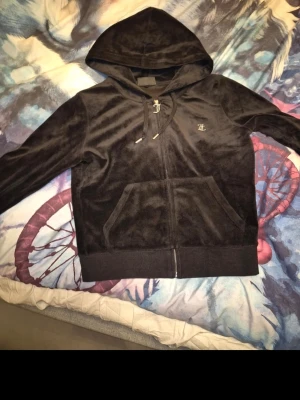 Svart hoodie från Juicy Couture - Mjuk och lyxig svart velour hoodie från Juicy Couture med dragkedja framtill, fickor och huva. Har snygga detaljer som JC-logga i strass på bröstet och metalländar på snörena. Perfekt för en chill och trendig look.