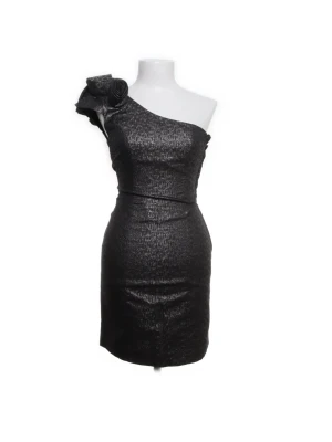 Svart one shoulder kortklänning - Snygg svart kortklänning med glittrig yta och one shoulder-design. Klänningen har en dekorativ blomdetalj på axeln och figurnära passform. Perfekt för dig som vill sticka ut med en elegant och trendig look.