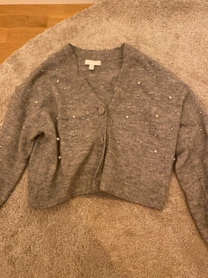 Grå kofta med pärlor - Säljer en grå kofta med pärldetaljer på både framsidan och längs ärmarna. Koftan har en knappar framtill, och är stickad i mjukt material. 