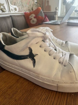 Vita sneakers med svart detalj - Snygga vita sneakers med svart detalj på sidan och hälen. Skorna har klassisk låg profil, vita skosnören och är tillverkade i slätt läder. Perfekta för en clean och stilren look. Skorna är från Axel Arigato och köptes för 2.200kr, defekter förekommer så priset går absolut att diskuteras!