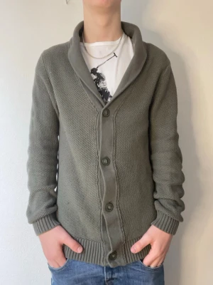 Olivgrön Cardigan från Lindbergh - Snygg olivgrön cardigan från Lindbergh. Defekterna visas på bild 5,6 och 7. Storlek medium och skicket är bra. Modellen på bilderna är 178cm och väger ca 70kg. Hör gärna av er vid funderingar eller frågor!